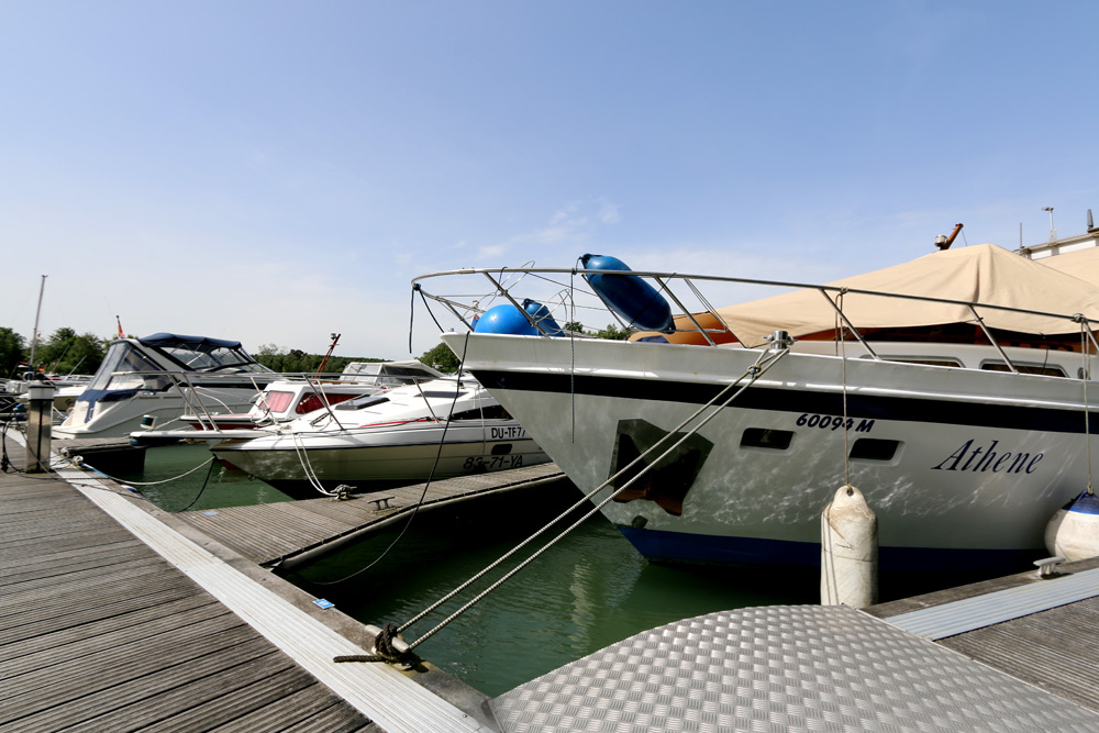 An einem sonnigen Tag liegen mehrere Motorboote in einem Yachthafen vor Anker, wobei ein Boot mit dem Namen Athene im Vordergrund zu sehen ist.