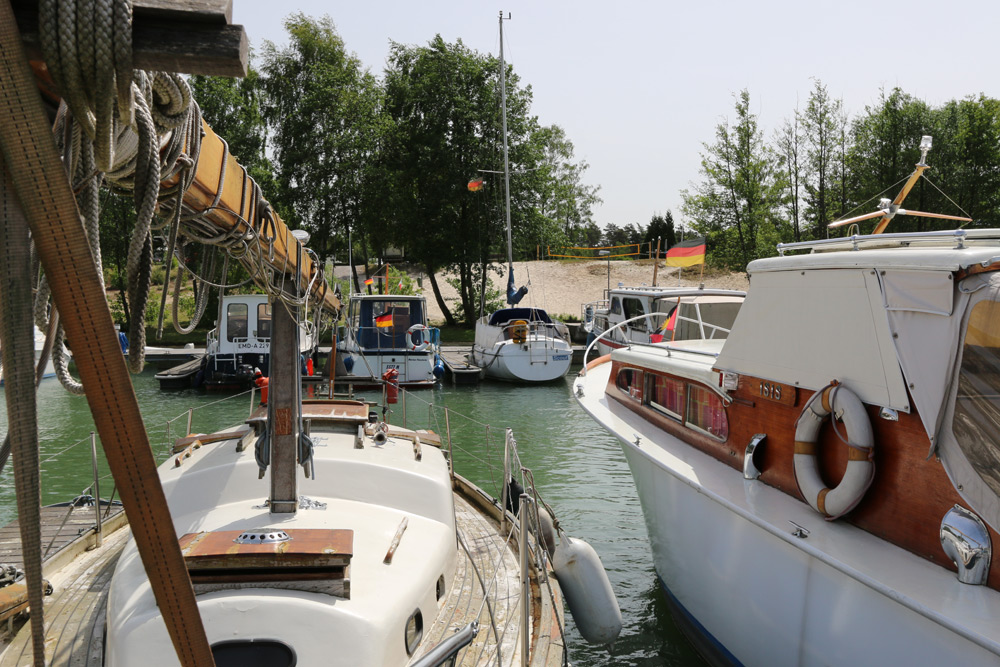 Mehrere Boote liegen in einem kleinen Yachthafen in der Nähe eines sandigen Ufers mit Bäumen; auf mehreren Booten sind deutsche Flaggen zu sehen.