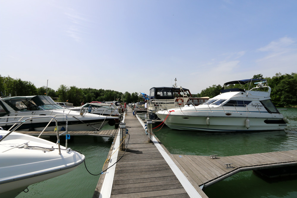 Mehrere Motorboote und Jachten liegen an einem Holzsteg an einem ruhigen, grünlichen Gewässer, umgeben von Bäumen und unter einem klaren Himmel.