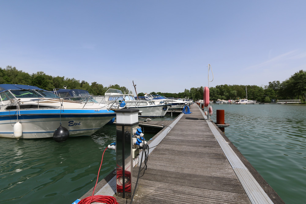 Mehrere Boote liegen an einem hölzernen Steg an einem ruhigen Gewässer, mit Bäumen und Grünpflanzen im Hintergrund unter einem klaren Himmel.