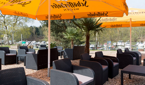 Sitzbereich im Freien mit schwarzen Korbstühlen und Tischen unter großen orangefarbenen Schöfferhofer-Sonnenschirmen, mit Blick auf einen Yachthafen mit Booten und umgeben von Bäumen.