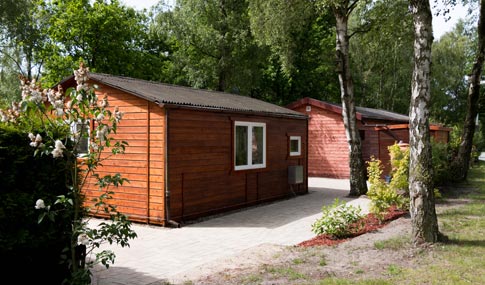 Zwei kleine Holzhütten stehen nebeneinander an einem gepflasterten Weg, umgeben von grünen Bäumen und Sträuchern in einer natürlichen Umgebung.