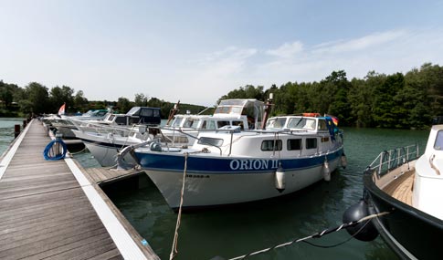 Mehrere Motorboote, darunter ein Boot mit dem Namen ORION II, liegen an einer Anlegestelle an einem ruhigen Gewässer mit Bäumen im Hintergrund unter einem klaren Himmel.