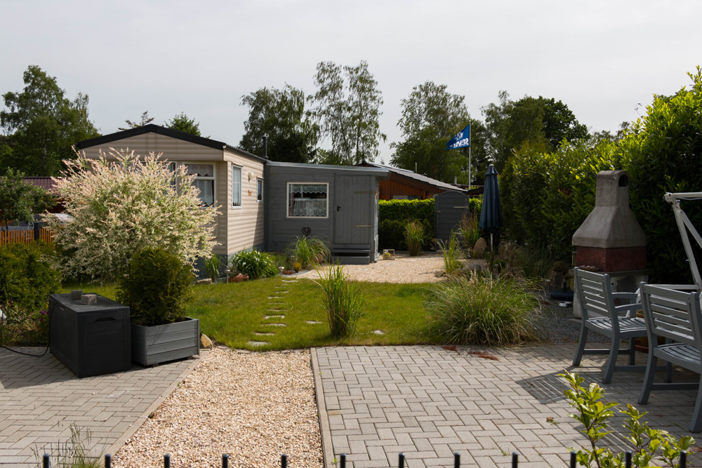 Ein kleiner Hinterhof mit einer gepflasterten Terrasse, einem Essbereich im Freien, einem Grill, Rasen, Sträuchern und einem hellen Gartenhaus inmitten von Grünflächen.