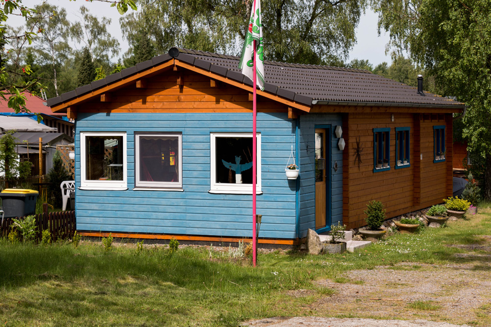 Ein kleines Holzhaus mit blauer Verkleidung und braunen Zierleisten steht auf einem grasbewachsenen Grundstück, mit Topfpflanzen am Eingang und einem Fahnenmast im Hof.