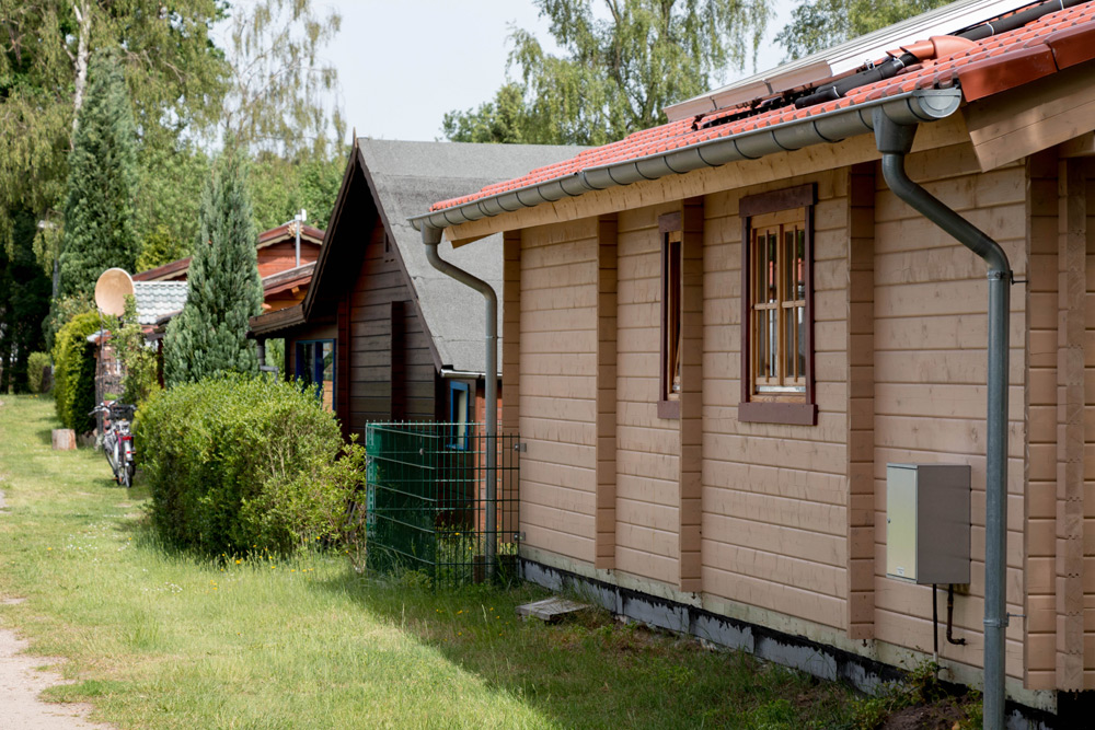Eine Reihe von kleinen Holzhäusern mit roten Ziegeldächern und Fenstern, umgeben von Grün und Bäumen an einem sonnigen Tag.