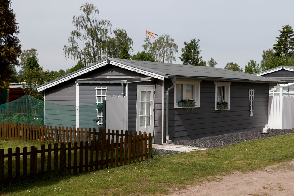 Ein kleines graues Holzhaus mit weißer Verkleidung, Blumenkästen an den Fenstern, einem Lattenzaun und einer Flagge auf dem Dach, umgeben von Gras und Bäumen.