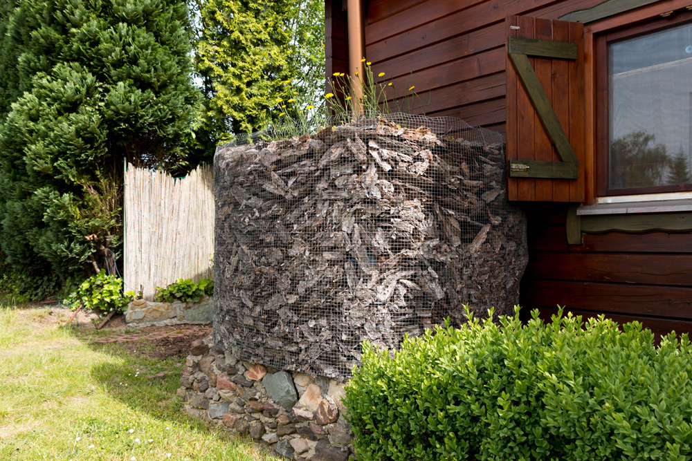 Eine mit Rinde und Steinen gefüllte Gabionenwand aus Drahtgeflecht steht neben einem Holzhaus mit einem offenen Fenster.