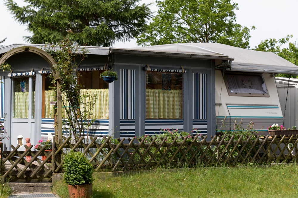 Ein gestreiftes Wohnmobil mit grünen und gelben Vorhängen ist hinter einem Holzzaun geparkt, umgeben von Pflanzen und Bäumen auf einem grasbewachsenen Grundstück.