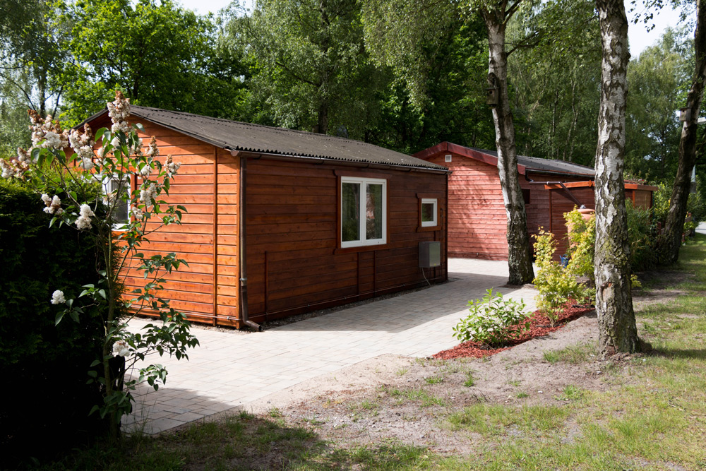Zwei Holzhütten mit Fenstern und kleinen Veranden befinden sich inmitten von Bäumen, gepflasterten Wegen und Grünflächen.