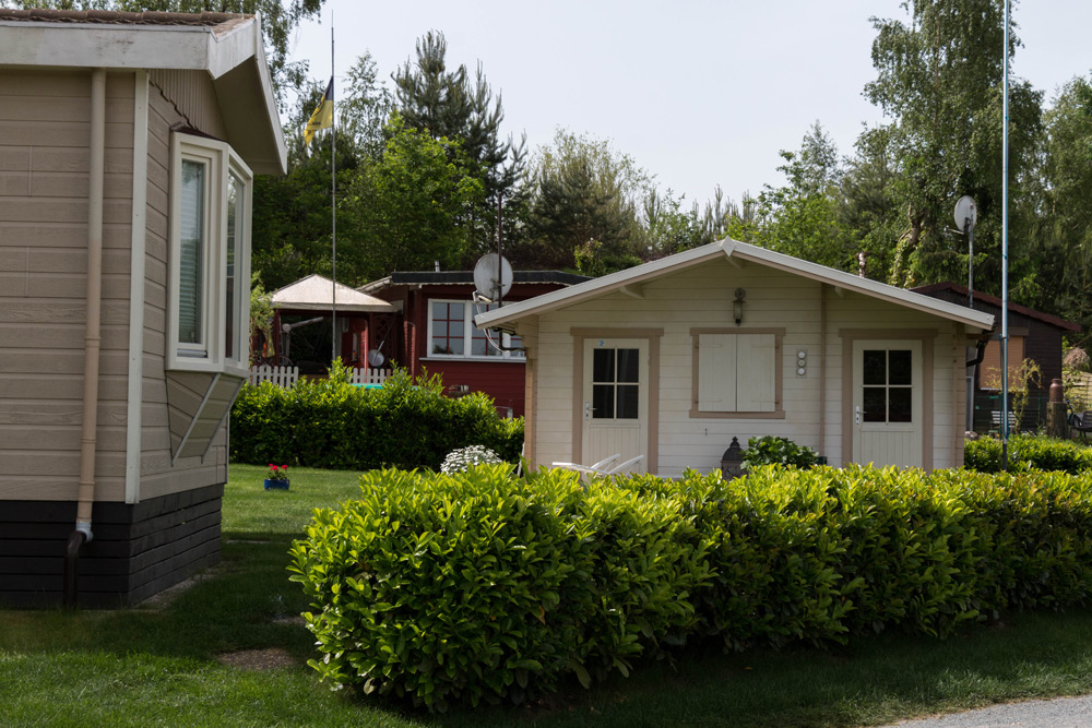 Ein kleines weißes Häuschen mit geschlossenen Fensterläden ist von grünen Sträuchern umgeben, während im Hintergrund auf einer Wiese andere Häuser und Bäume zu sehen sind.