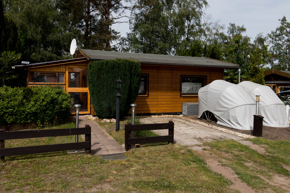 Ein kleines Holzhaus mit einer Satellitenschüssel auf dem Dach, eine Hecke davor und zwei überdachte Fahrzeuge, die neben einer Kiesauffahrt geparkt sind.