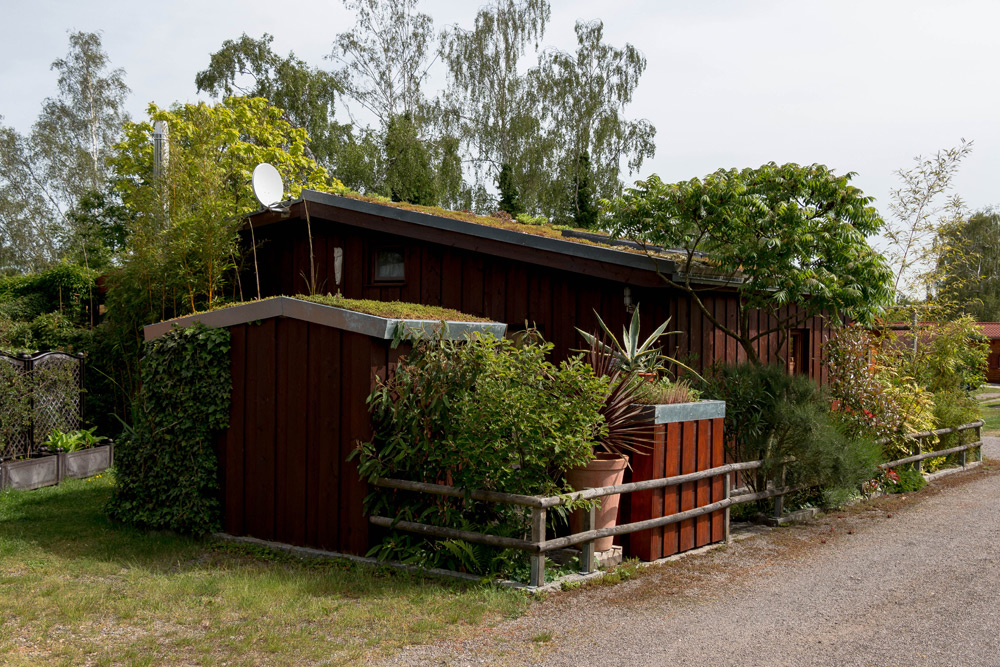 Ein kleines, rotes Holzhaus mit grünem Dach und Pflanzen drumherum, neben einem Kiesweg gelegen und von Bäumen und Sträuchern umgeben.