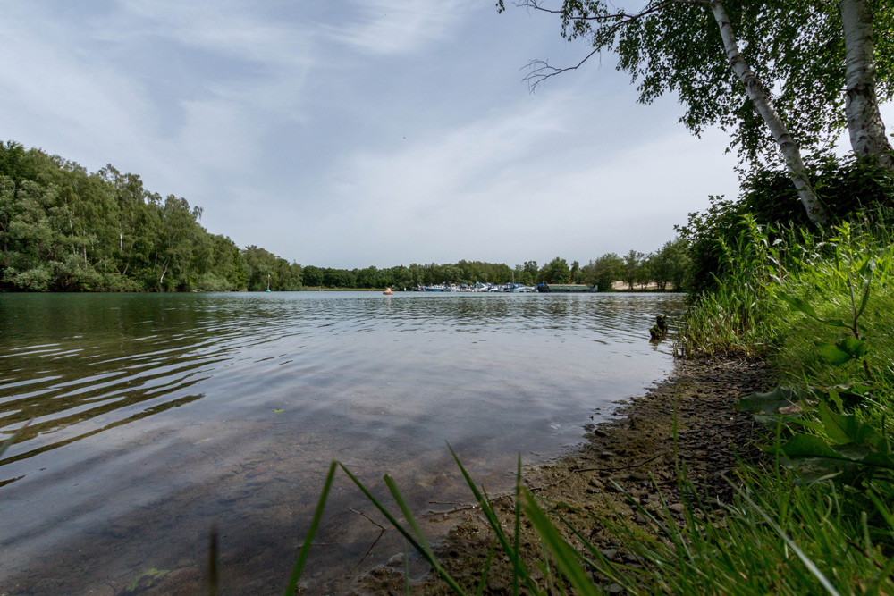 Ein ruhiger See mit klarem Wasser, Bäumen am Ufer und Booten in der Ferne unter einem bewölkten Himmel.