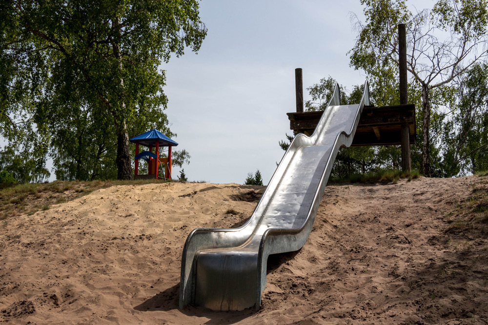 Auf einem Spielplatz im Freien ist eine Metallrutsche in einen Sandhügel gebaut. Im Hintergrund sind Bäume und ein blau überdachter Unterstand zu sehen.