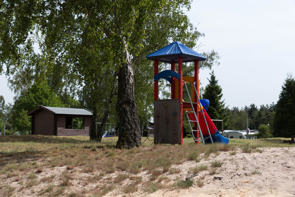 Ein Spielplatz mit Rutsche, Kletterwand und Leiter steht auf sandigem Boden in der Nähe von Bäumen und einer kleinen Holzhütte.