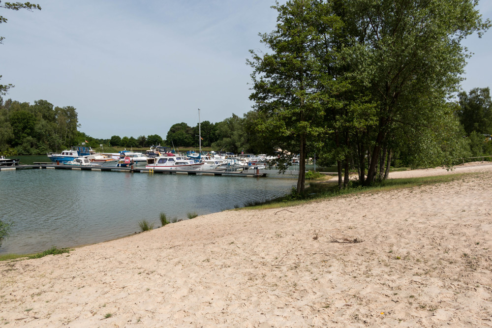 Ein Sandstrand führt zu einem kleinen Jachthafen mit mehreren angedockten Booten, gesäumt von Bäumen und ruhigem Wasser unter einem teilweise bewölkten Himmel.