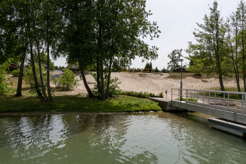 Eine kleine Brücke überquert ein ruhiges Gewässer und führt zu einer sandigen Fläche mit Bäumen und spärlichem Gras im Hintergrund.