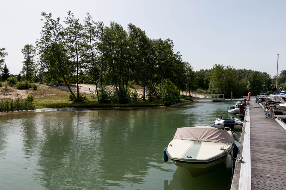 Ein angedocktes Motorboot liegt an einem hölzernen Steg auf einem ruhigen, grünlichen Fluss mit Bäumen und sandigen Ufern im Hintergrund unter einem klaren Himmel.