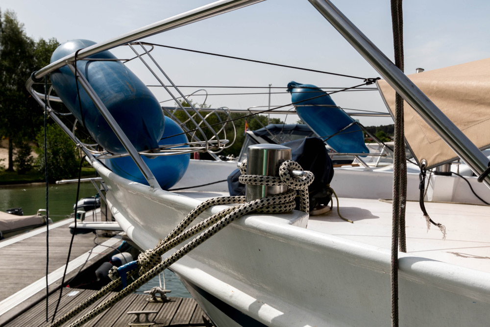 Nahaufnahme des Bugs eines angedockten Bootes mit Seilen, die an einer Metallklampe befestigt sind, blauen Fender, die an der Reling angebracht sind, und einem Steg, der neben dem Boot zu sehen ist.