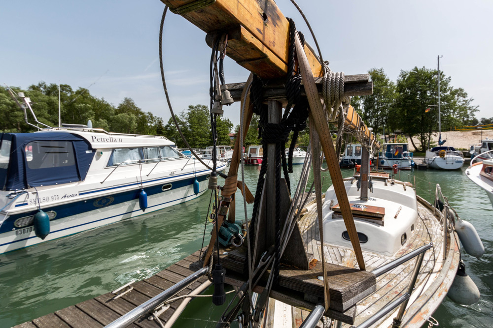Eine Nahaufnahme eines hölzernen Segelbootmastes mit Takelage, der an einem sonnigen Tag in einem Jachthafen angedockt ist, umgeben von anderen Booten und grünem Wasser.