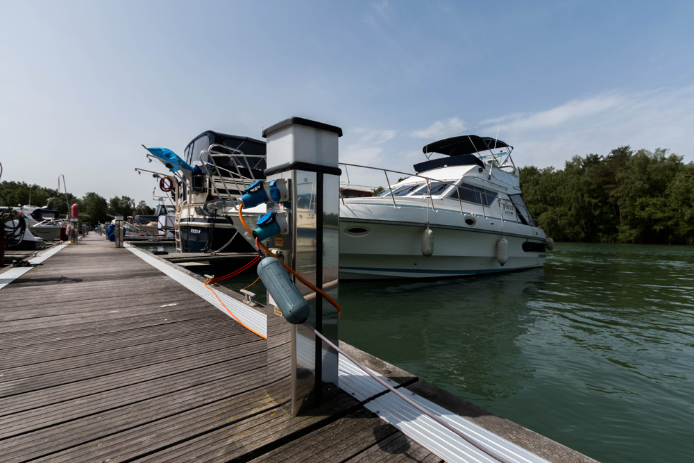 Eine Motoryacht liegt in einem Jachthafen an einem Holzsteg vor Anker. An einem klaren Tag sind im Hintergrund Bäume und andere Boote zu sehen.