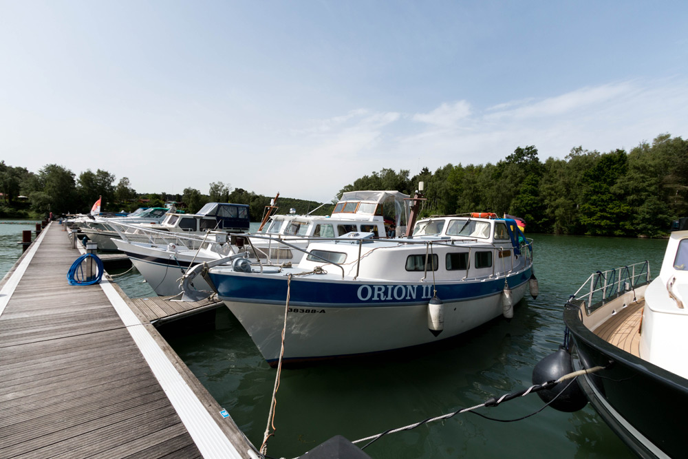 Mehrere Boote, darunter eines mit dem Namen ORION II, liegen an einem hölzernen Steg an einem See, mit Bäumen und Grünpflanzen im Hintergrund und einem teilweise bewölkten Himmel.