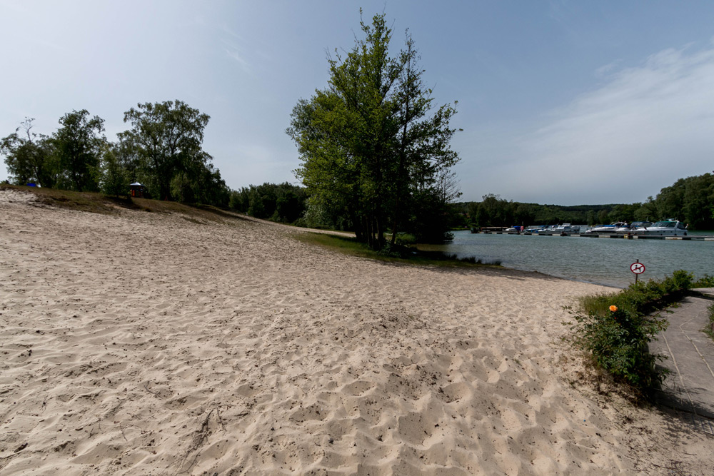 Sandstrand mit Bäumen am Rande eines Gewässers, in der Ferne angedockte Boote und ein kleiner Weg auf der rechten Seite.