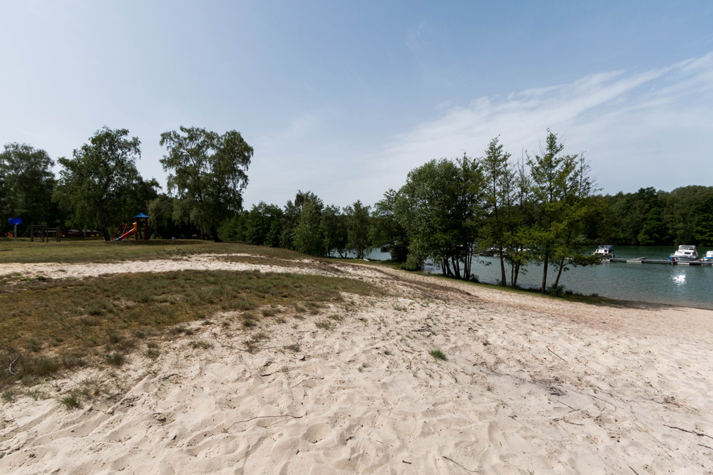 Sandstrand mit spärlichem Gras, Bäumen im Hintergrund und Booten auf einem Fluss unter einem teilweise bewölkten Himmel.