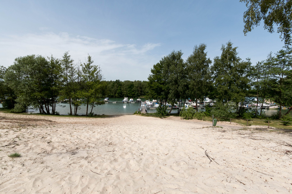 Ein Sandweg führt zu einem kleinen Jachthafen mit Booten, die an einem baumbestandenen Ufer unter einem teilweise bewölkten Himmel ankern.