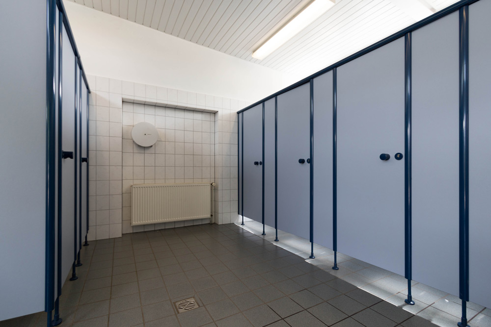 Eine Reihe geschlossener blauer Toilettenkabinen mit gefliesten Wänden und Böden, einem Heizkörper und einer runden Wandleuchte unter Leuchtstoffröhren.