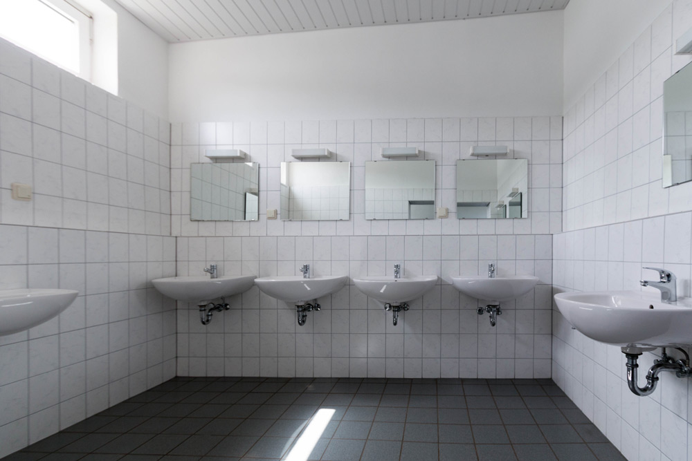 Eine Reihe von fünf weißen Waschbecken mit Spiegeln über ihnen in einer gefliesten öffentlichen Toilette mit grauen Bodenfliesen und weißen Wänden.