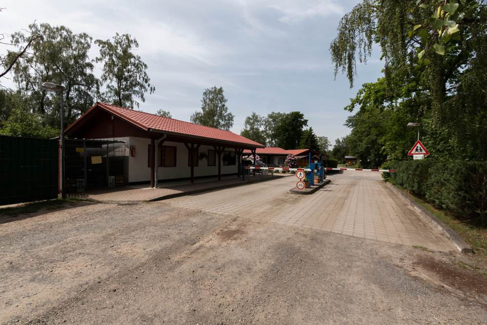 Ein kleines Gebäude mit rotem Dach neben einer Toreinfahrt mit Verkehrsschildern und Schranken, umgeben von Bäumen unter einem teilweise bewölkten Himmel.