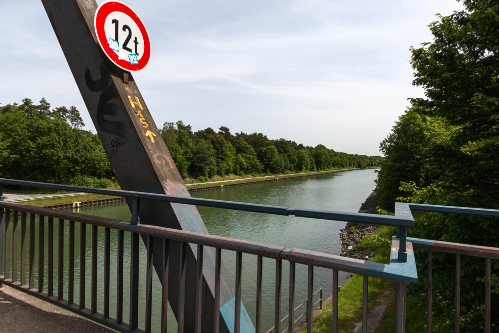 Blick von einer Brücke über einen von Bäumen gesäumten Kanal, mit einem 12-Tonnen-Schild und Metallgeländern im Vordergrund.
