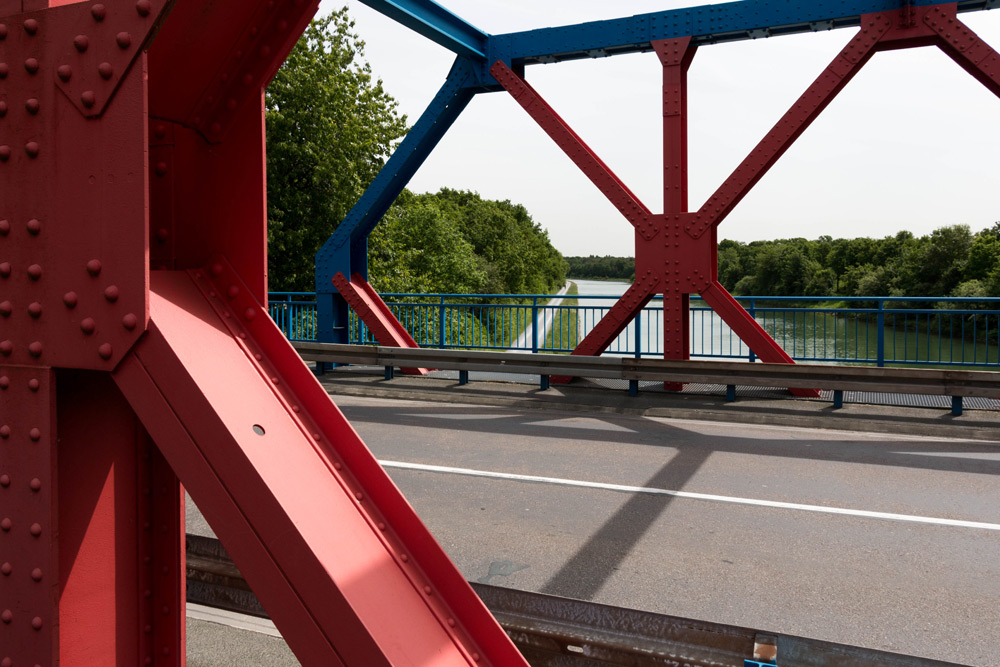 Rote und blaue Stahlbrückenstruktur mit einer Straße, mit Blick auf einen Fluss und grüne Bäume im Hintergrund.