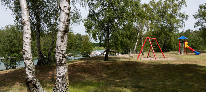 Spielplatz mit roten Schaukeln und einer Rutsche neben einem See, umgeben von grünen Bäumen und Grasflächen.