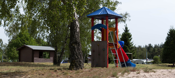 Ein Spielplatz mit einem blau überdachten Spielgerät, einer Kletterwand und einer roten Rutsche befindet sich auf sandigem Boden in der Nähe von Bäumen und einer kleinen Holzhütte.