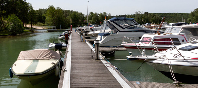 Holzsteg mit mehreren Motorbooten und Yachten auf beiden Seiten in einem ruhigen, von Bäumen gesäumten Yachthafen.