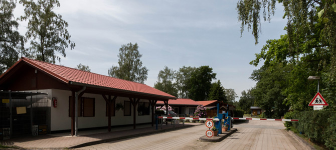 Ein kleines Gebäude mit rotem Dach steht neben einer geschlossenen Schranke auf einer gepflasterten Zufahrt, umgeben von Bäumen; Verkehrs- und Fußgängerschilder sind sichtbar.