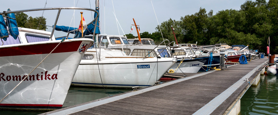 Mehrere kleine Motorboote liegen an einem Holzsteg an einem ruhigen, von Bäumen gesäumten Gewässer vor Anker. Die Bootsnamen Romantika, Scout und Lauren sind sichtbar.
