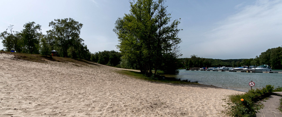 Ein Sandstrand mit Bäumen auf der linken Seite und ein Gewässer mit mehreren angedockten Booten auf der rechten Seite unter einem klaren Himmel.