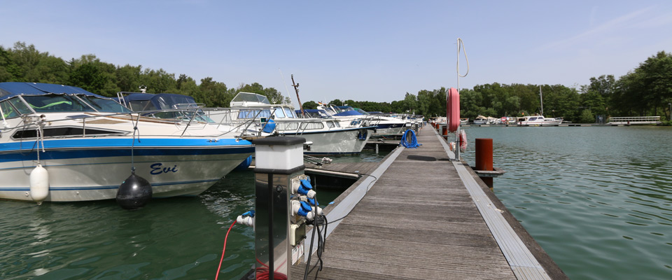 Ein hölzerner Steg erstreckt sich über das Wasser mit mehreren vertäuten Motorbooten auf der linken Seite und Bäumen im Hintergrund unter einem klaren Himmel.