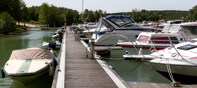 Mehrere Motorboote und Yachten liegen an einem hölzernen Steg in einem ruhigen Jachthafen, umgeben von Bäumen und Grünpflanzen unter einem klaren Himmel.