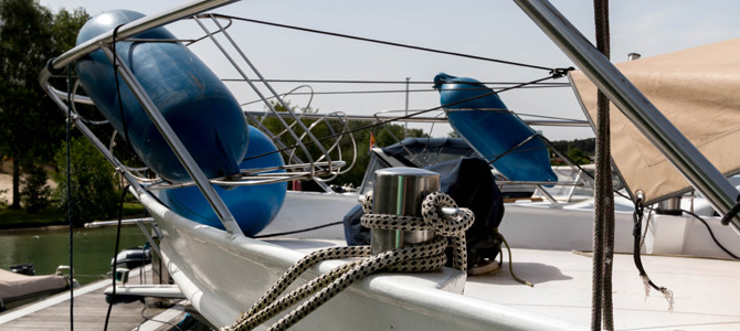Ein Seil, das um eine Metallklampe auf dem Deck eines angedockten Bootes gewickelt ist, mit blauen Fendern, die an der Reling befestigt sind.