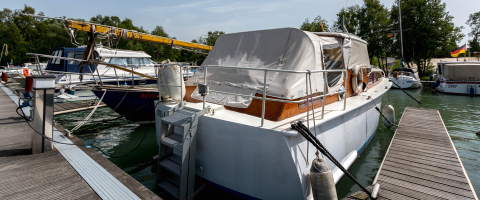 Ein überdachtes Motorboot liegt in einem Yachthafen neben anderen Booten, im Hintergrund sind Bäume und eine Sandfläche zu sehen.