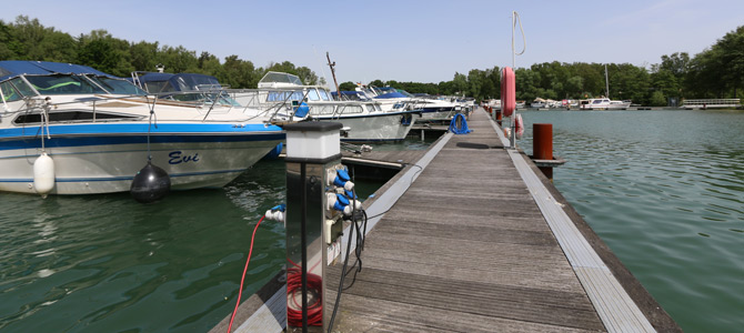 Holzsteg mit festgemachten Booten auf der linken Seite, Wasser auf der rechten Seite, Versorgungspfosten und Bäume im Hintergrund unter einem klaren Himmel.