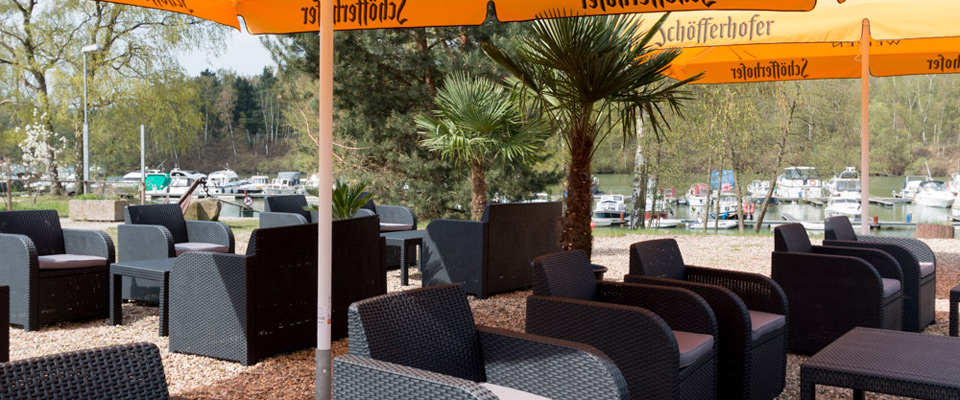 Außensitzbereich mit schwarzen Korbstühlen und Tischen unter großen gelben Sonnenschirmen, mit Blick auf einen Yachthafen mit Booten und umgeben von Bäumen.