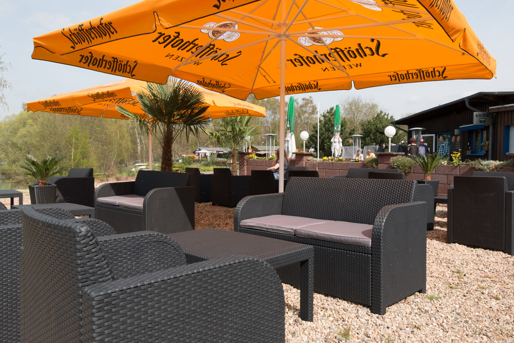 Sitzbereich im Freien mit schwarzen Korbstühlen und Tischen unter großen orangefarbenen Sonnenschirmen auf einer Kiesfläche, mit Pflanzen und einem Gebäude im Hintergrund.
