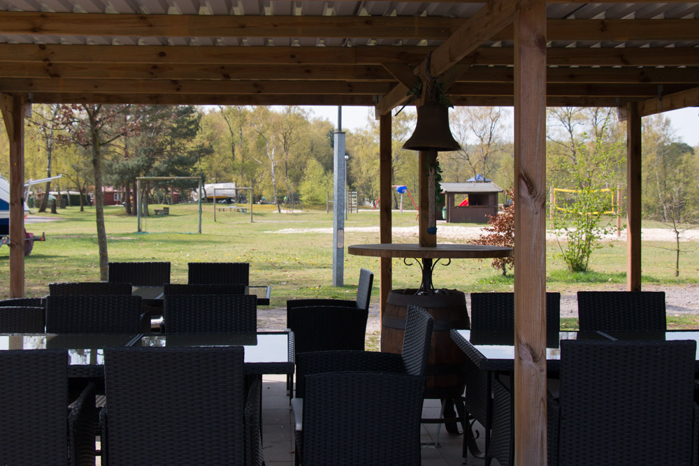 Sitzbereich im Freien mit schwarzen Korbstühlen und Glastischen unter einer hölzernen Pergola mit Blick auf einen grasbewachsenen Park mit Bäumen und Spielgeräten im Hintergrund.