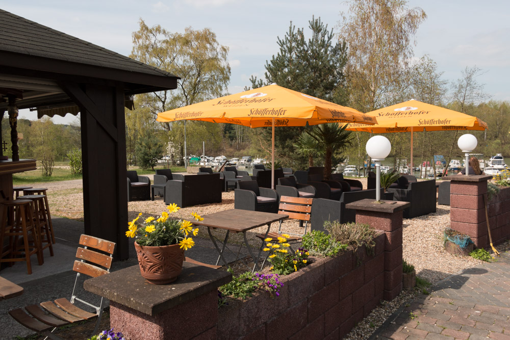Sitzbereich im Freien mit schwarzen Stühlen und gelben Sonnenschirmen, Holztischen, Topfblumen und einem Blick auf geparkte Boote und Bäume im Hintergrund.